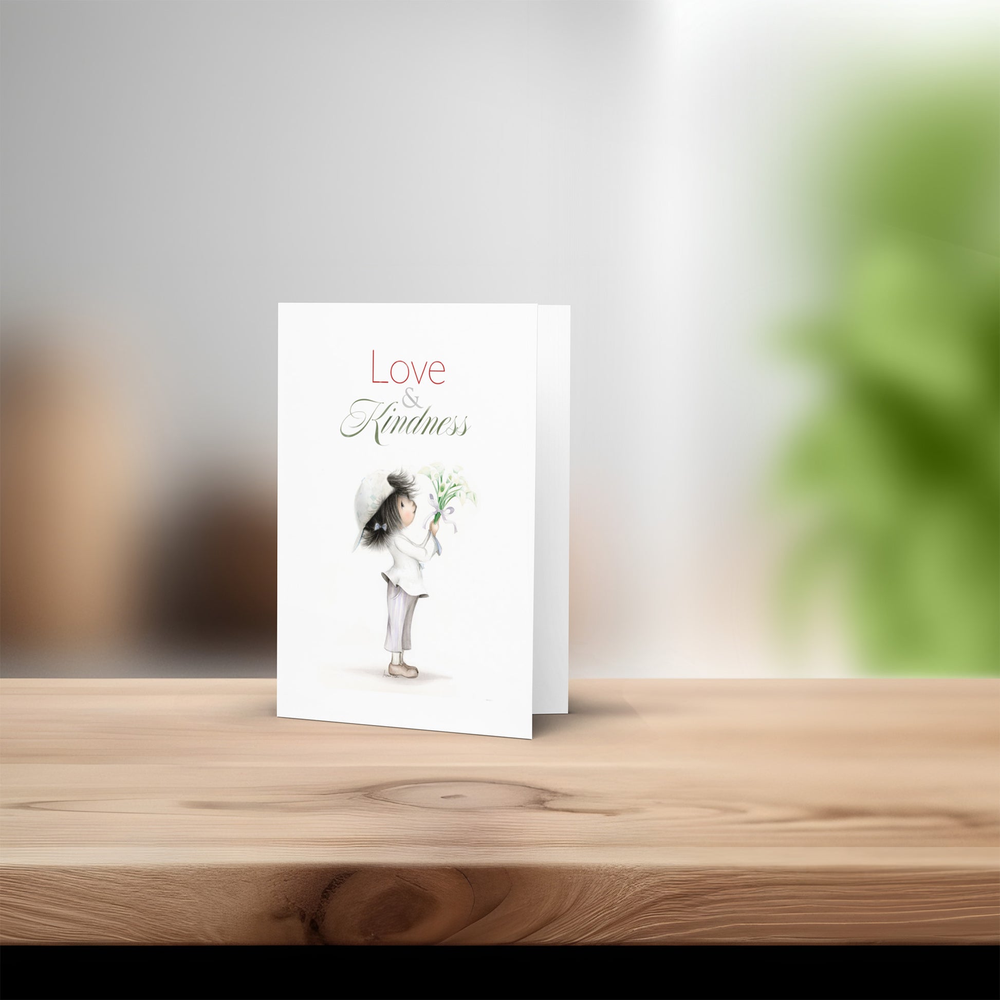 Greeting card with a child illustration and 'Love Kindness' text on a wooden surface.