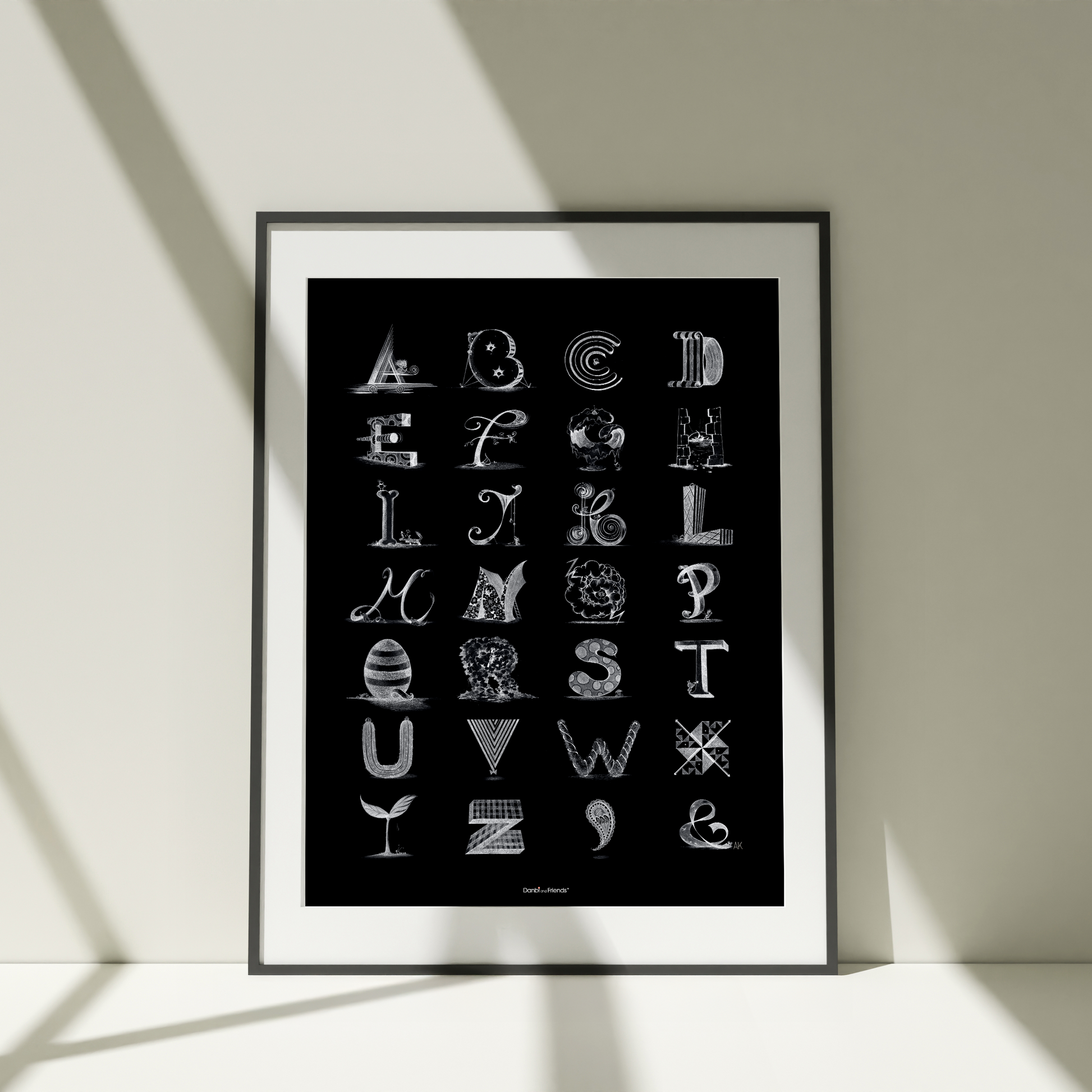 Framed poster with stylized alphabet letters on a black background, leaning against a light-colored wall.