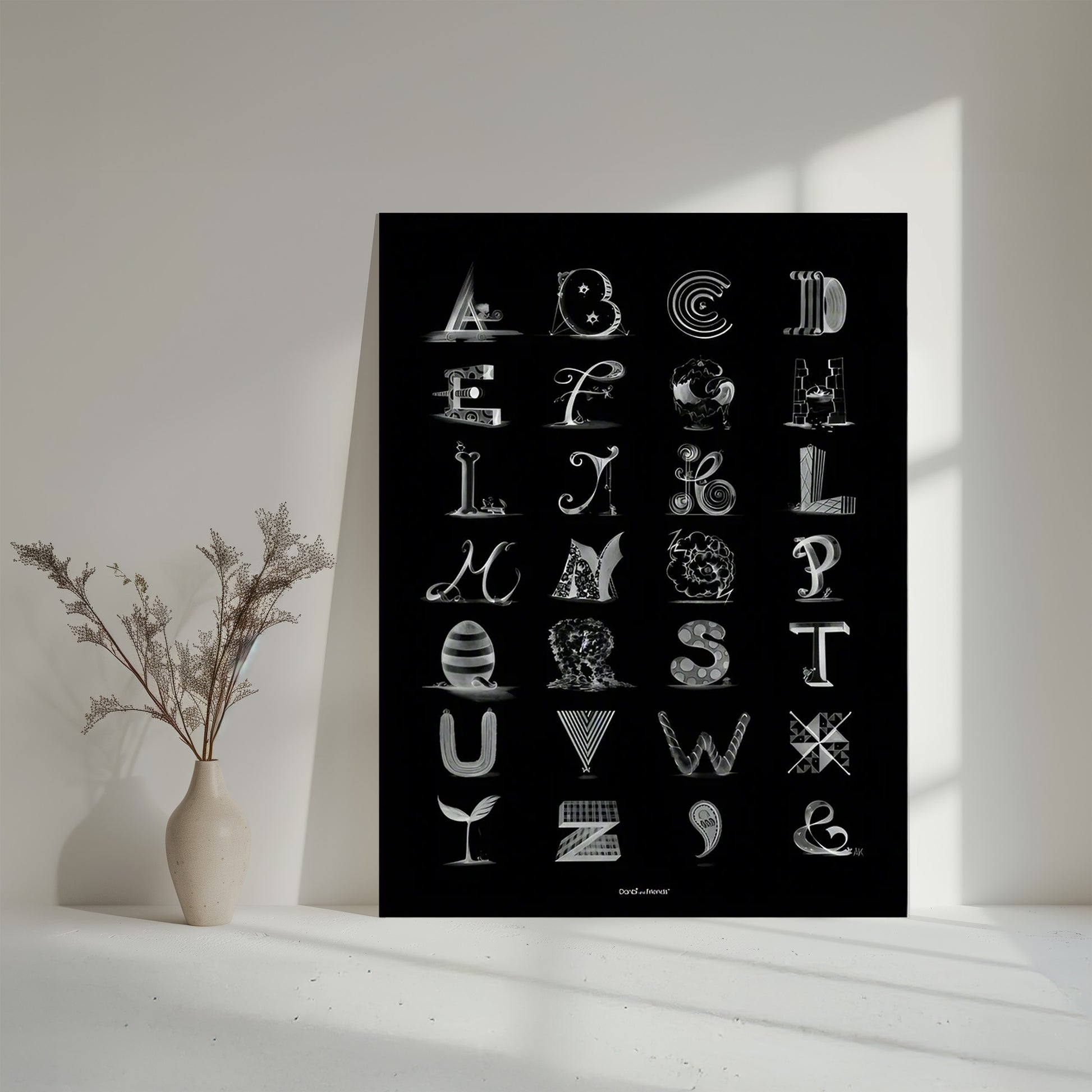 Black poster with white alphabet design on a white surface with a vase of dried flowers.