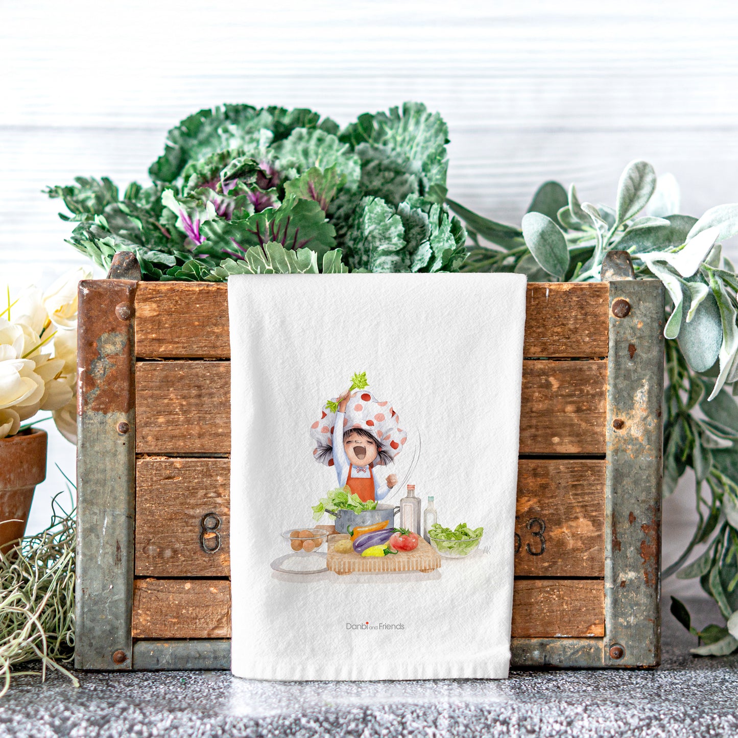 Tea towel with a kitchen-themed design in front of a wooden crate with plants