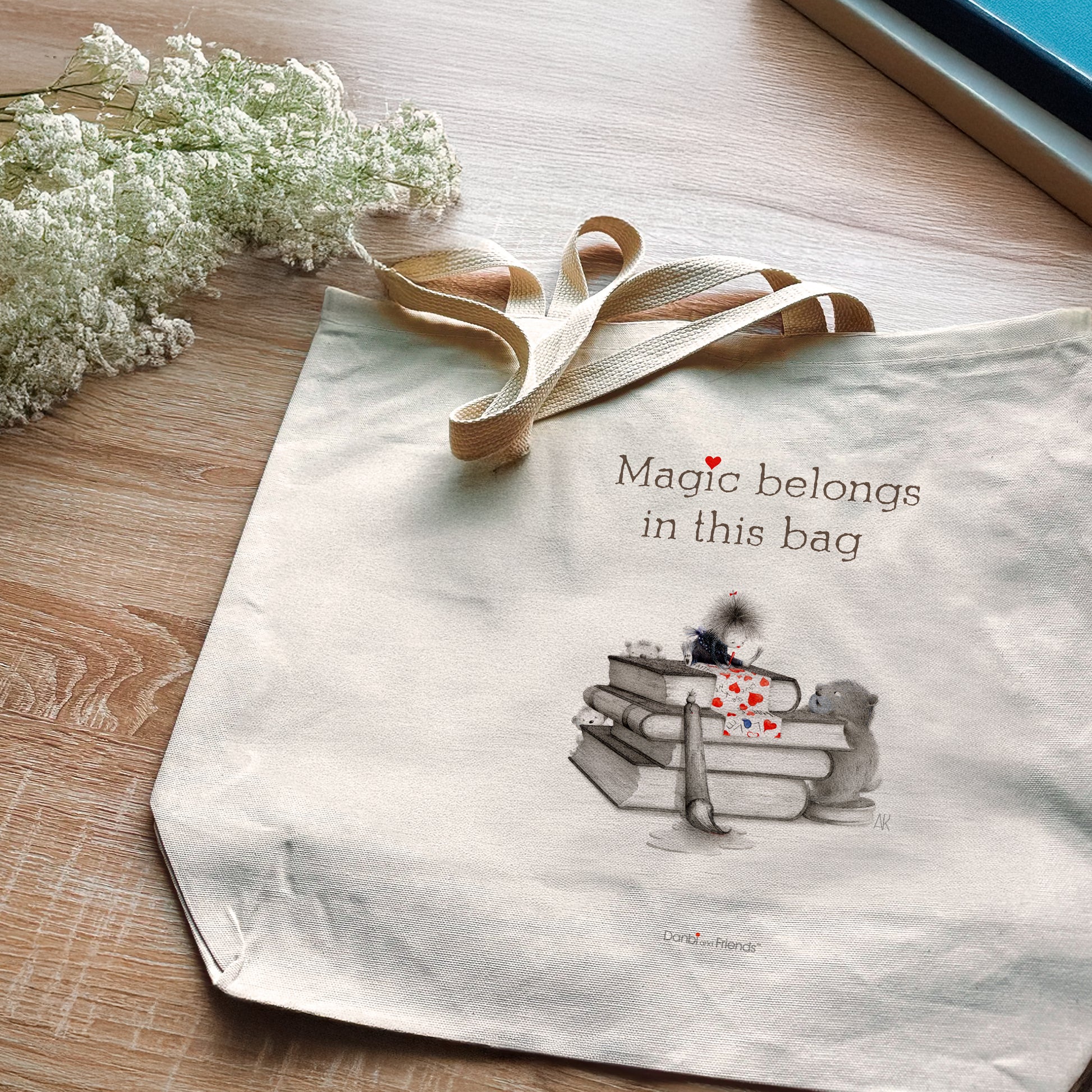 Tote bag with 'Magic belongs in this bag' text and a graphic of books and a heart on a wooden surface with flowers.