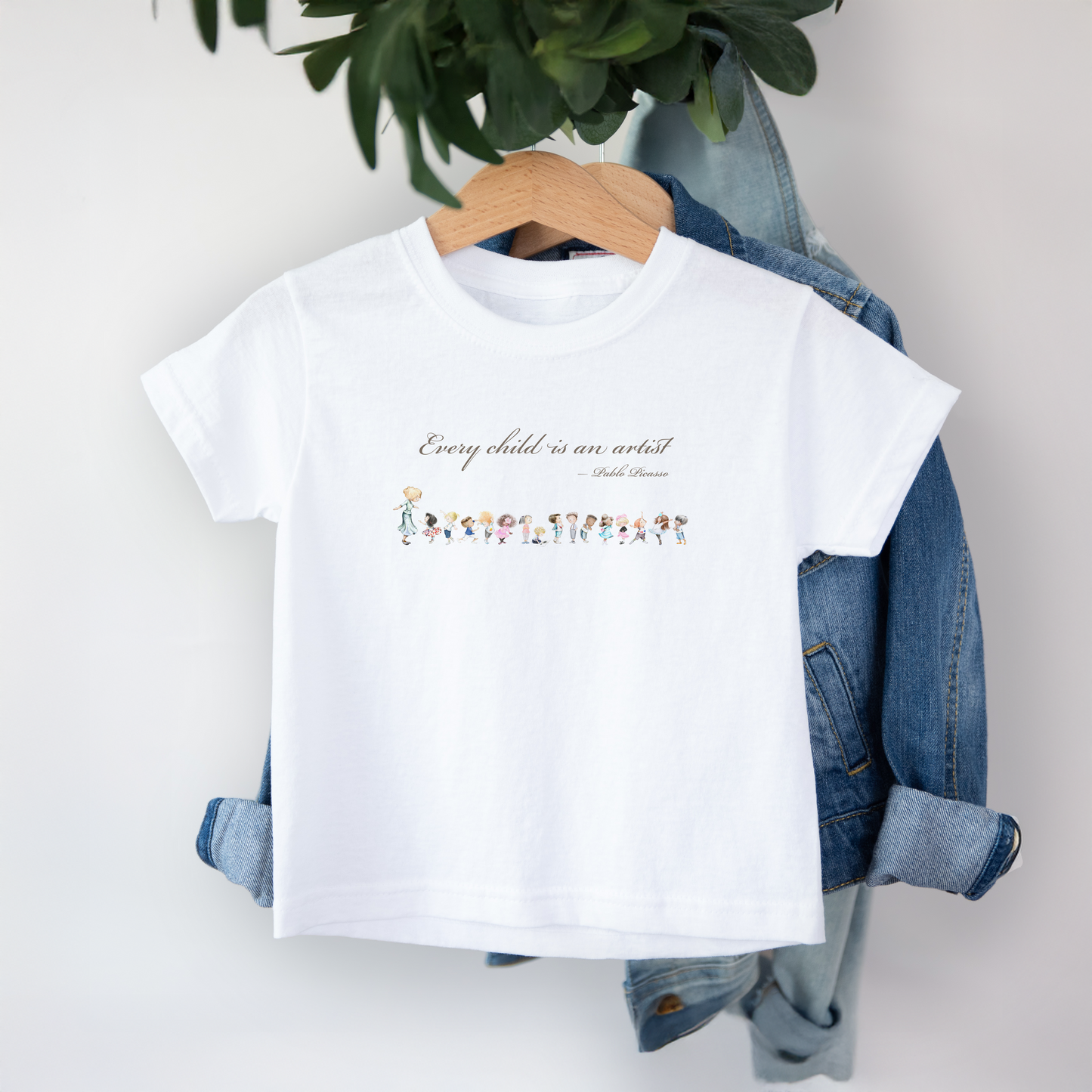 White t-shirt with text and graphics on a hanger with denim jacket and plant in the background